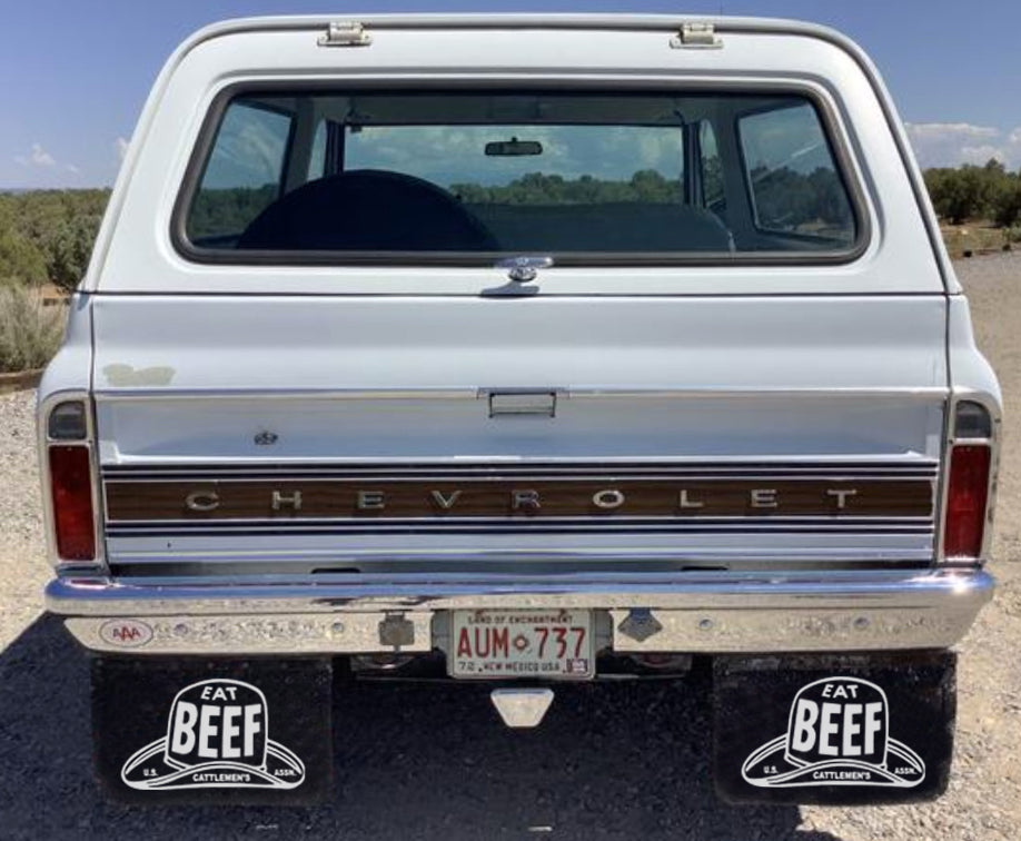 Back view of a silver Chevrolet truck with 'Eat Beef' mudflaps on a desert road.