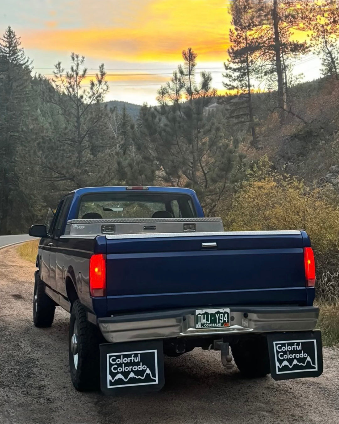 Blue pickup truck with retro Colorado state mudflaps installed on a road with trees and sunset in the background