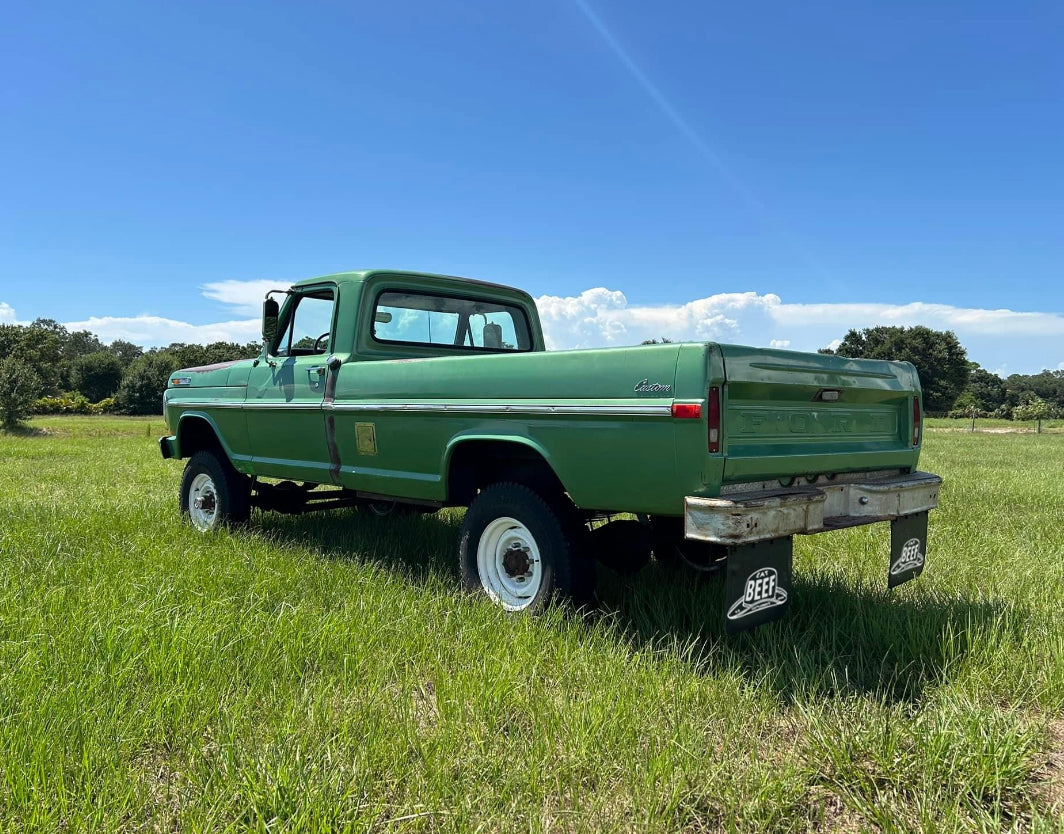 Green pickup truck parked on grass with a clear blue sky and Eat Beef mudflaps installed